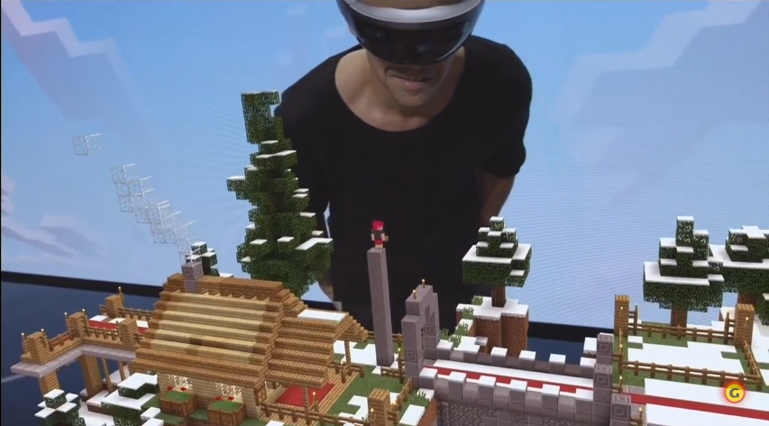 Picture of a man wearing the HoloLens on his head gazing down at a hologram of a Minecraft world. He sees his tiny friend standing on a tower in the game.